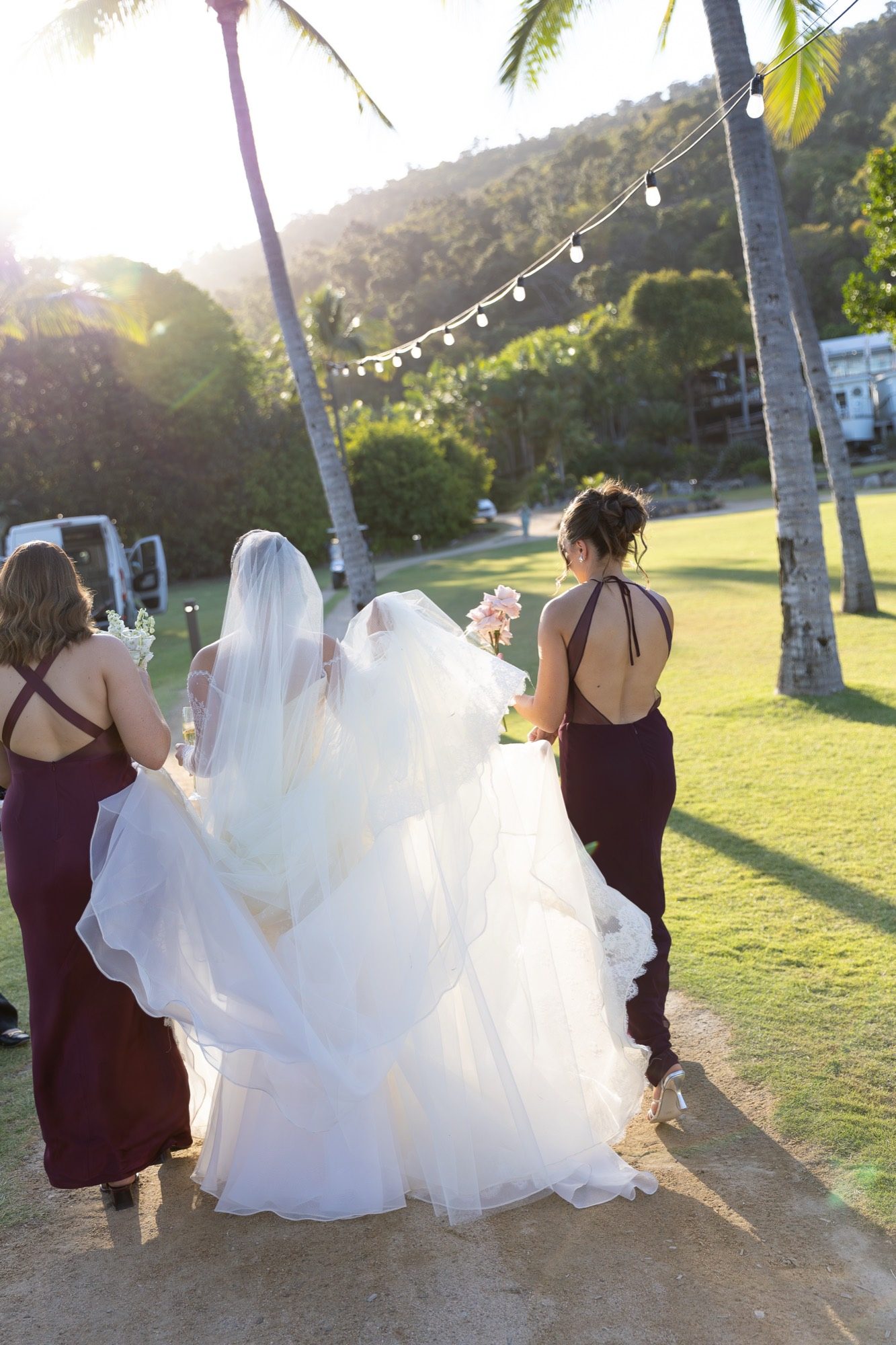 Bridesmaids carrying the bride's train at golden hour beneath festoon lights and palms, Freedom Shores, Airlie Beach